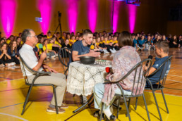 Štirje ljudje sedijo okoli okrogle mize, prekrite z belo čipkasto prtjo, na športnem parketu. Na mizi je črna skleda in nekaj predmetov. Eden od udeležencev drži mikrofon, drugi ima pred seboj roke, kot da nekaj razlaga. V ozadju sedi večja skupina ljudi, večinoma oblečenih v rumene majice, ob steni osvetljeni z rožnatimi reflektorji. Celoten dogodek poteka v telovadnici, kar nakazujejo lesena tla in športne oznake.
