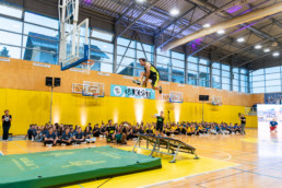 športni dvorani poteka akrobatski nastop pred množico gledalcev. Na sredini slike je akrobat v črno-rumeni športni opremi, ki med skokom z mini trampolina leti proti košarkarskemu obroču. Pod trampolinom je zelena varnostna blazina. V ozadju sedi skupina ljudi ob rumeni steni, nad katero visi transparent z napisom »38. Krst«. Na steni so nameščeni košarkarski koši, prostor pa je osvetljen z reflektorji in naravno svetlobo skozi velika okna.