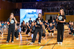 Skupina mladih nastopa v športni dvorani. V ospredju stojijo trije posamezniki z mikrofoni, eden drži tudi list papirja. V ozadju sedi in kleči večja skupina ljudi, vsi oblečeni v temne majice z belim potiskom. Na levi strani je velik zaslon, ki prikazuje notranjost prostora, dvorana pa je osvetljena z močno umetno svetlobo.