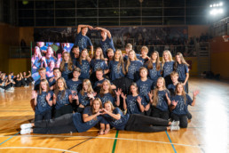 Skupinska fotografija več kot dvajsetih oseb, oblečenih v enotne temno modre majice z belim potiskom, ki prikazuje številko »4B«. Osebe so postavljene v več vrst na leseni športni dvorani, nekatere klečijo, druge stojijo, dve osebi na vrhu oblikujeta srce z rokami. V ozadju je vidna tribuna z občinstvom in velik zaslon, ki prikazuje podoben prizor. Osvetlitev je močna, usmerjena na skupino.