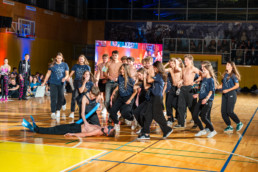 Skupina mladih nastopa na odru v športni dvorani med plesno točko. Večina nastopajočih je oblečena v temne hlače in majice z napisi, nekateri so zgoraj brez. Na tleh leži oseba, ki je del koreografije, medtem ko drugi stojijo okoli nje v dinamičnih pozah. V ozadju je velika LED zaslonska tabla z barvitimi grafikami, za njo pa občinstvo, ki spremlja dogajanje. Osvetlitev je živahna, z modrimi in oranžnimi reflektorji, kar ustvarja energično atmosfero.