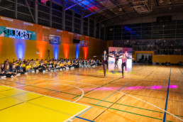 Notranjost športne dvorane med prireditvijo. Na sredini parketa trije nastopajoči izvajajo plesno točko, osvetljeni z barvnimi reflektorji. V ozadju sedi večja skupina ljudi ob steni, razdeljena v dve skupini – ena v rumenih majicah, druga v temnih oblačilih. Na steni visi transparent z napisom »38. KRST«. Dvorana je osvetljena z modrimi, rdečimi in oranžnimi lučmi, nad parketom so vidni košarkarski obroči in konstrukcija stropa.