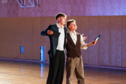 Dve osebi stojita na leseni športni dvorani, oblečeni v elegantna oblačila. Ena oseba nosi temen suknjič in drži črn pravokoten predmet, druga oseba ima bel srajco in rjav telovnik ter drži tablico. Obe osebi imata roke rahlo razprte, kot da nekaj predstavljata ali razlagata. V ozadju je lesena stena in del košarkarskega obroča.