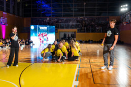 Skupina mladih v rumenih majicah sedi v krogu na parketu športne dvorane. Na levi strani stoji oseba v črni majici z mikrofonom in listom v roki, na desni pa druga oseba v črni majici drži rumen predmet. V ozadju je velik LED zaslon z barvno grafiko in napisom, za njim pa tribune z občinstvom v zatemnjeni dvorani. Na tleh so vidne črte igrišča za košarko, prostor je osvetljen z močnimi reflektorji.