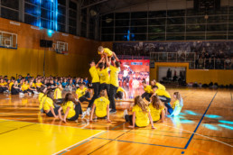 Skupina mladih v rumenih majicah izvaja skupinsko akrobatsko točko v športni dvorani. Nekateri sedijo na tleh, medtem ko več članov dviguje osebo nad glavo. V ozadju so gledalci, športni rekviziti in velika LED zaslonska tabla z osvetlitvijo. Dvorana ima lesena tla, košarkarske koše in rumene stene.