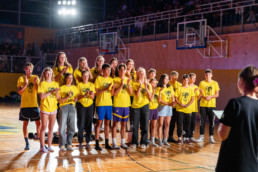 Skupina mladih stoji na športnem igrišču v telovadnici, oblečeni v enotne rumene majice z napisom »1.C« in ilustracijo sove. Osebe so postavljene v vrsto, nekatere nosijo športne hlače, druge kratke hlače ali dolge hlače. V ozadju so vidni košarkarski koši, tribune z občinstvom in močne reflektorske luči, ki osvetljujejo prizorišče. Na desni strani slike je oseba, ki drži papir, kot da vodi dogodek.