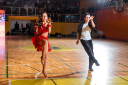 Plesni par izvaja energičen latinskoameriški ples na parketu športne dvorane. Oseba na levi nosi rdečo obleko z resicami in plesnimi čevlji z visoko peto, medtem ko oseba na desni nosi črno jakno, belo majico in črne hlače. V ozadju so vidne tribune z občinstvom in velika LED-zaslonka, ki prikazuje dogajanje.