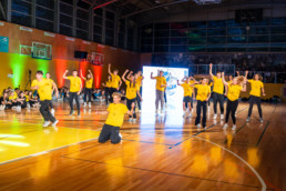 Skupina mladih nastopa na odru v športni dvorani. Vsi nosijo rumene majice in temne hlače ter izvajajo usklajen plesni nastop na leseni košarkarski površini. V ozadju so gledalci, barvne luči (rdeča, zelena) osvetljujejo steno, na sredini pa je velik svetlobni zaslon z grafiko.