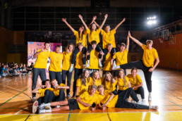 Skupina mladih v rumenih majicah pozira v telovadnici, postavljeni v večplastno piramido. Nekateri stojijo, drugi klečijo ali sedijo na tleh, eden na desni strani drži nogo v zraku v gimnastični pozi. V ozadju so vidni gledalci, velika LED-zaslon in športna oprema, prostor je osvetljen z močnimi reflektorji.