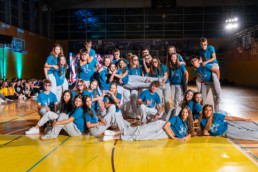 Skupina ljudi v športni dvorani pozira za skupinsko fotografijo. Vsi nosijo enake modre majice in sive hlače, kar nakazuje, da gre za plesno ali športno ekipo. Nekateri člani stojijo, drugi klečijo ali ležijo na tleh, medtem ko enega člana dvigujejo v vodoravni položaj. V ozadju so vidni gledalci, športna oprema in osvetlitev, ki ustvarja živahno vzdušje dogodka.