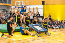 Skupina nastopajočih izvaja akrobatsko točko v telovadnici pred občinstvom. Na sredini je oseba, ki sedi na ramenih druge osebe in drži košarkarsko žogo, medtem ko več drugih članov ekipe sodeluje pri dvigu in podpiranju. Na desni strani je postavljen trampolin, v ozadju pa so gledalci, ki sedijo na tleh in na tribunah. Na steni so rumene in lesene obloge, v prostoru pa je športna oprema, kot so blazine in gimnastične letve.