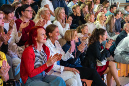 Skupina ljudi sedi na lesenih tribunah v notranjem prostoru, videti je kot dvorana ali prireditveni prostor. Osebe so obrnjene proti odru ali dogajanju izven kadra in ploskajo. Večina nosi priložnostna oblačila v svetlih in nevtralnih barvah, med njimi izstopa oseba v rdečem puloverju s pikami. V ospredju so torbice in jakne, ozadje je zapolnjeno z več vrstami sedečih ljudi.