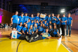 Skupinska fotografija večje skupine mladih, ki sedijo in stojijo na športnem parketu v telovadnici. Vsi nosijo enake modre majice z belim logotipom na sprednji strani. Nekateri držijo plakate in listke, eden prikazuje ilustracijo živali, drugi fotografijo. V ozadju so vidne tribune, osvetlitev z reflektorji ter druge skupine ljudi, ki sedijo ob robu prostora.