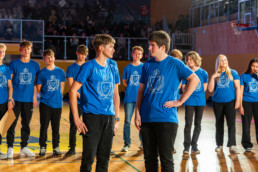 Skupina ljudi stoji v športni dvorani na lesenem parketu. Vsi nosijo enake modre majice z belim grbom in napisom na sprednji strani. Dva posameznika v ospredju se pogovarjata, medtem ko ostali stojijo v ozadju v vrsti. V ozadju so vidne tribune z občinstvom in mreža ob igrišču. Osvetlitev je močna, usmerjena na skupino na sredini dvorane.