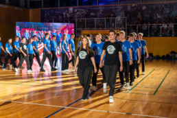 Dijaki prvih letnikov v športni dvorani prihajajo na prizorišče, da bodo povedali svečano prisego. Večina nosi modre majice z belim logotipom, nekaj jih ima rumene, mentorji pa črne majice z enakim motivom. Nastop poteka na parketni površini z označenimi črtami, v ozadju pa je velika LED zaslonska stena z živahnimi barvami in tribune z gledalci