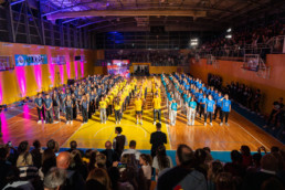 Vsi razredi prvošolcev stojijo v vrstah na športnem igrišču v telovadnici. Oblečeni so v enotne majice modrih in rumenih odtenkov. V ospredju je občinstvo, ki sedi in stoji, medtem ko opazuje dogajanje. V ozadju so osvetljene stene z rožnato in vijolično svetlobo ter transparent z napisom »KRIST«. Na desni strani so tribune polne gledalcev, nad igriščem pa je kovinska konstrukcija strehe z reflektorji.