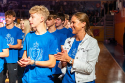 Skupina ljudi stoji v notranjem prostoru na športnem parketu, oblečeni v enake modre majice z belim grbom, krono in lovorovim vencem na sprednji strani. Ena oseba v ospredju nosi čez majico svetel jeans jakno. Osebe ploskajo, v ozadju so vidne tribune z občinstvom in košarkarski obroč. Osvetlitev je močna in poudarja ljudi v ospredju.