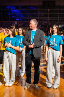 Skupina ljudi stoji v športni dvorani na parketnem podu. V ospredju je oseba v temnem suknjiču in športnih copatih, obkrožena z osebami v modrih majicah z belim potiskom in sivimi trenirkami. V ozadju so vidni drugi udeleženci v rumenih in modrih majicah ter osvetljena tribuna z modro-vijolično osvetlitvijo.