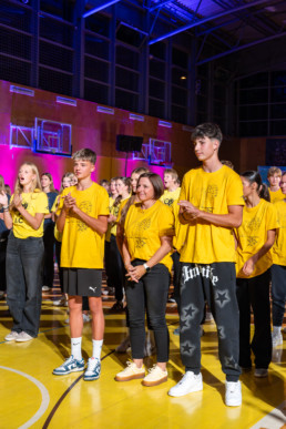 Skupina mladih stoji v športni dvorani, oblečeni v enotne rumene majice z natisnjenim motivom na sprednji strani. Nekateri nosijo športne copate, drugi casual obutev. Osebe so postavljene v vrsto in gledajo proti isti smeri, kot da spremljajo dogodek ali nastop. V ozadju so vidne športne naprave, kot so košarkarski koši, ter stena z lesenimi oblogami. Prostor je osvetljen z barvnimi reflektorji, ki oddajajo rožnato in modro svetlobo, kar ustvarja živahno vzdušje.