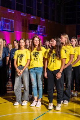 Skupina mladih stoji v telovadnici na parketu, oblečeni v rumene majice z napisom »1.C« in grafiko, ki vključuje športne simbole. V ozadju so vidni košarkarski obroči in stena telovadnice, osvetljena z rožnato in vijolično scensko svetlobo. Za njimi stojijo osebe v modrih majicah, kar nakazuje na organiziran dogodek, verjetno šolski sprejem ali krst.