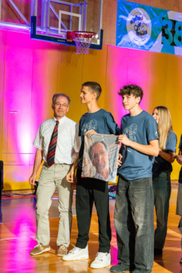 Na fotografiji je skupina ljudi v modrih majicah, ki stoji na športnem parketu v telovadnici. Dva posameznika v ospredju držita nad glavo kos blaga z natisnjeno podobo moškega obraza. V ozadju je velik transparent z napisom »38. KRST« in dodatnim besedilom »PRVOSOLOV«, ob njem pa logotip. Na levi strani je košarkarski obroč, prostor pa je osvetljen z rožnato in modro scensko svetlobo.