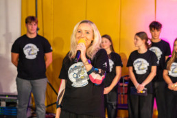 Oseba stoji v ospredju in drži mikrofon, oblečena v črno majico z velikim potiskom, ki prikazuje ilustracijo z napisom Čas teče, Šentvid ostaja. To je ravnateljica šole, ki pozdravlja prvošolce.V ozadju stoji več ljudi, prav tako oblečenih v enake črne majice z istim potiskom. Prostor ima rumeno steno in je osvetljen z odrskimi lučmi, na desni strani je opazna rožnata svetloba.