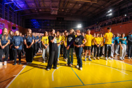 Velika skupina ljudi stoji v športni dvorani z leseno podlago in visoko streho. Osebe so razporejene v več vrst, oblečene v majice različnih barv – modre, rumene in črne –, kar nakazuje na pripadnost različnim skupinam ali ekipam. V ospredju je nekaj ljudi, ki se pogovarjajo, medtem ko drugi stojijo z rokami ob telesu ali prekrižanimi. V ozadju so vidni košarkarski obroči, tribune in močna osvetlitev, ki osvetljuje prostor. Na levi strani je opazna vijolična osvetlitev, kar daje dogodku prazničen videz. Celotna scena nakazuje na šolski dogodek oz. družabno srečanje.