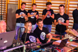 Skupina ljudi sedi in stoji v prostoru z lesenimi letvami na steni. V ospredju sta dve osebi za mizo z računalniško opremo, kabli in mešalno konzolo. Na mizi so tudi prenosni računalnik, mikrofon in radijska postaja. V ozadju stoji več oseb, ki nosijo enake črne majice z velikim grafičnim logotipom na sprednji strani, nekatere imajo roke sklenjene ali ploskajo. Osvetlitev je topla, kar daje vtis dogodka ali tehnične priprave