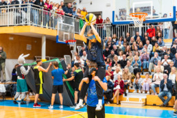 Na sliki je športna prireditev v telovadnici, kjer je veliko gledalcev na tribunah. V ospredju oseba sedi na ramenih druge osebe, obe imata prevezo čez oči. Oseba zgoraj drži košarkarsko žogo nad glavo, pripravljena na met proti košu. V ozadju so drugi udeleženci v športnih oblačilih in maskota, ki stoji ob zloženi blazini. Na tribunah ljudje opazujejo dogajanje.