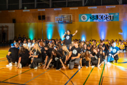 Skupina mladih v črnih majicah z belim logotipom izvaja koreografijo na športnem parketu v telovadnici. V ospredju so plesalci v različnih pozah, eden stoji na sredini z dvignjeno roko in mikrofon v drugi roki. V ozadju sedi večja skupina ljudi, osvetljena z modrimi in zelenimi reflektorji. Na steni visi transparent z napisom '38. KRST' in logotipom dogodka