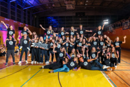 Velika skupina mladih stoji v športni dvorani na svetlem parketu, oblečeni so v enake črne majice z belim logotipom na sprednji strani. Skupina je razporejena v več vrst, nekateri stojijo, drugi klečijo ali ležijo na tleh, nekaj jih dviguje druge v zrak. V ozadju so vidne tribune, košarkarski koši in osvetljena dvorana z visokim stropom. Vsi imajo dvignjene roke ali pozirajo v igrivih položajih, kar daje vtis veselja in timskega duha.