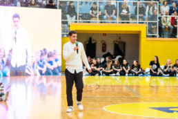 Oseba stoji na sredini športne dvorane in drži mikrofon, kot da govori občinstvu. Na tleh je leseni parket z rumenimi in modrimi oznakami igrišča. V ozadju sedi skupina ljudi ob steni, večinoma oblečenih v črna oblačila z belimi logotipi na majicah. Za njimi je rumena stena in mrežasta ograja, za katero so gledalci na tribunah. Na levi strani slike je velik zaslon, ki prikazuje povečano sliko govorca.