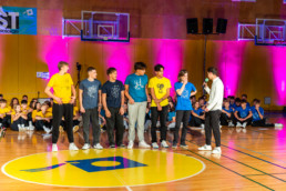 Skupina šestih oseb stoji na sredini športne dvorane na rumenem krogu z modrim logotipom. Osebe nosijo majice različnih barv, predvsem rumene, modre in temne, z natisnjenimi motivi. Ena oseba na desni drži mikrofon in govori skupini. V ozadju sedi večja skupina ljudi, večinoma v rumenih in modrih majicah, ob steni osvetljeni z rožnatimi lučmi. Na steni so vidni košarkarski obroči in transparent z napisom KRST