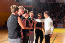 Skupina mladih stoji v vrsti v športni dvorani, oblečeni so v črna oblačila brez rokavov. Oseba v belem puloverju drži mikrofon in ga ponuja enemu izmed nastopajočih. V ozadju so vidne tribune, polne gledalcev, ki spremljajo dogodek. Na tleh je parket z označenimi črtami za športne igre.