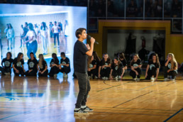 Oseba, ki poje stoji na sredini športne dvorane in drži mikrofon, kot da govori ali poje. V ozadju sedi skupina ljudi v črnih majicah z belimi logotipi, razporejenih v vrsti ob robu igrišča. Za njimi je velik zaslon, na katerem je prikazana slika več oseb, ki stojijo in se pogovarjajo. Tla so lesena, z označenimi črtami za športne igre, prostor pa je osvetljen z močno umetno svetlobo.