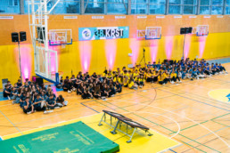 Skupina dijakov sedi na tleh športne dvorane, razdeljena v več skupin po barvah majic (temno sive, rumene in modre). V ozadju je rumena stena z več košarkarskimi koši, nad katerimi visi transparent z napisom »38. KRST«. Na steni so nameščeni reflektorji, ki oddajajo rožnato svetlobo. V ospredju je trampolin in zelena blazina za telovadbo.