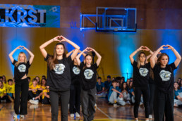 Skupina nastopajočih izvaja plesno točko v telovadnici. V ospredju je šest oseb, oblečenih v črne majice z belim potiskom in črne hlače, ki z rokami oblikujejo srce nad glavo. V ozadju sedi večja skupina gledalcev ob steni, ki je osvetljena z modrimi reflektorji. Na levi strani je vidna velika modra in bela transparenta z napisom »KRST PRVOSOLCEV«, nad sredino pa košarkarski obroč in konstrukcija.