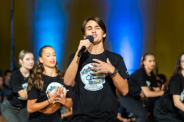 Skupina ljudi izvaja plesno točko na odru z modro osvetlitvijo v ozadju. Oseba v ospredju drži mikrofon in nosi črno majico z grafiko, ki prikazuje logotip dogodka ali šole. Ostali nastopajoči so oblečeni v podobne majice in so postavljeni v različnih plesnih pozah. Na fotografiji je pevec Anže Strajnar.