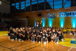 Skupina mladih ljudi izvaja plesno točko v športni dvorani. Večina nastopajočih je oblečena v črna oblačila z belimi športnimi copati, nekateri imajo na rokah srebrne trakove. V ospredju so plesalci v počepu, medtem ko oseba na sredini stoji in drži mikrofon z dvignjeno roko. V ozadju so gledalci, ki sedijo na tribunah, ter skupina ljudi v rumenih majicah. Na steni visi transparent z napisom »38. KR« in logotipom, prostor pa je osvetljen z modrimi reflektorji.