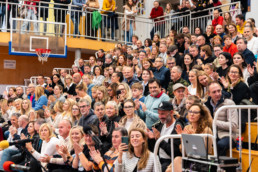 Velika skupina ljudi sedi na tribunah v športni dvorani. V ospredju so vidne več vrst sedežev, kjer ljudje sedijo tesno skupaj in ploskajo. V ozadju je zgornja galerija z dodatnimi gledalci, ki stojijo ob ograji. Na levi strani slike je košarkarski obroč z mrežico, nad njim oranžna stena. Na desni strani spodaj je vidna kamera na stojalu, usmerjena proti dogajanju. Barvna paleta vključuje oranžne, bele in kovinske odtenke, značilne za športne dvorane.