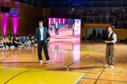 Notranjost športne dvorane z leseno parketno podlago, osvetljeno z močno odrsko svetlobo in vijoličnimi reflektorji ob steni. V ospredju stojita dve osebi na igrišču, oblečeni v elegantna oblačila, medtem ko v ozadju sedi skupina ljudi v vrstah ob steni, oblečeni v barvne majice. Na desni strani je velik LED zaslon, ki prikazuje odsev dogajanja na igrišču. V ozadju so vidni tribuni z dodatnimi gledalci in športnimi rekviziti.