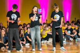 Tri osebe stojijo na odru telovadnice, oblečene v črne majice z belim logotipom in držijo mikrofone ter listke. V ozadju sedi večja skupina ljudi v rumenih in temno modrih majicah. Stena je osvetljena z rožnatimi in vijoličnimi lučmi, kar ustvarja živahno atmosfero dogodka.