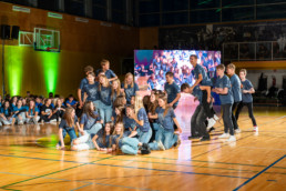 Skupina mladih stoji in sedi na parketu športne dvorane, oblečeni so v modre majice in kavbojke. V ospredju je več oseb, ki tvorijo zgoščeno skupino, nekatere klečijo ali sedijo, druge stojijo ob strani. V ozadju je večja skupina ljudi, ki sedi ob steni, ter velik zaslon, na katerem je prikazan posnetek dogajanja na odru. Dvorana ima lesena tla, visoke stene in osvetlitev z zelenimi reflektorji.