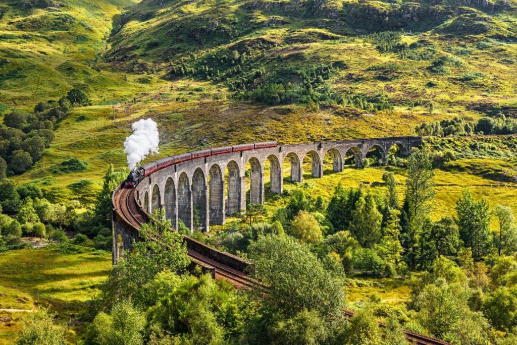Parna lokomotiva vozi po viaduktu Glenfinnan, obdanim z zelenimi škotskimi griči; prizorišče je znano iz filma Harry Potter, kjer vozi čarovniški vlak proti Bradavičarki.