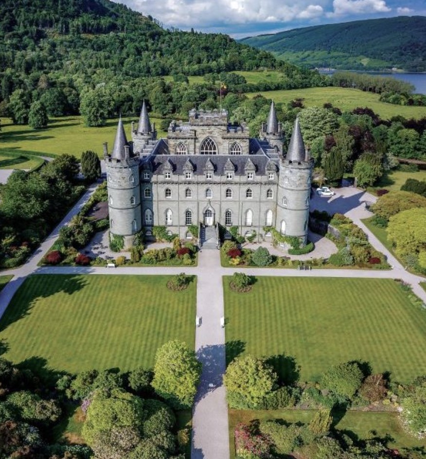 Zračni posnetek gradu Inveraray v škotskem okrožju Argyll and Bute s štirimi stolpi, obdanega z urejenimi vrtovi in zeleno pokrajino s hribi v ozadju.