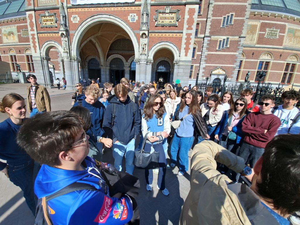 Skupina stoji pred okrašenim opečnatim poslopjem z obokanimi vhodi in dekorativnimi elementi. Stavba deluje zgodovinsko ali kulturno pomembna, verjetno gre za muzej ali znamenitost.