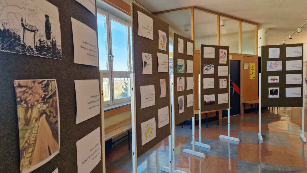 Razstava likovnih in literarnih del v šolskem hodniku. Na več oglasnih panojih so razstavljene risbe in listi z besedili, postavljeni vzdolž oken, skozi katera sije dnevna svetloba. Na tleh je pisan odsev parketa, v ozadju pa so vidna vrata in omarice.