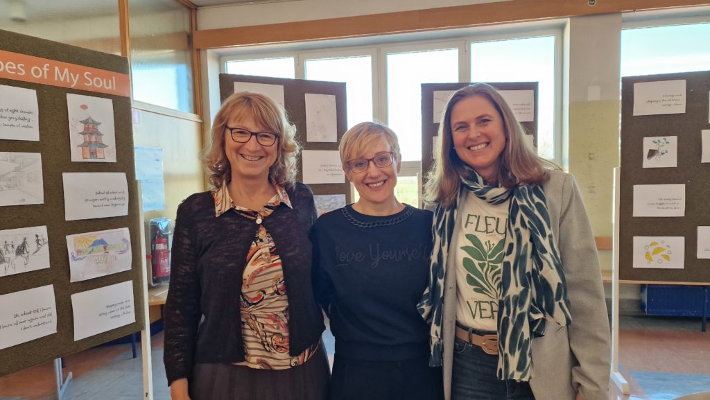 Tri profesorice angleščine stojijo nasmejane pred razstavnimi panoji z risbami in pesmimi v šolskem hodniku.