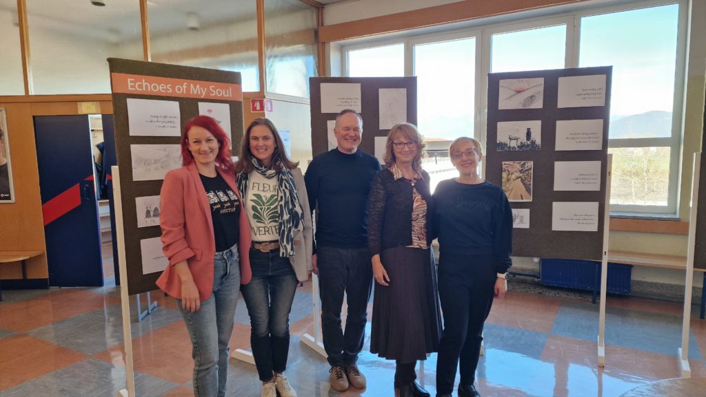 Vsi člani aktiva anglistov stojijo nasmejani pred razstavnimi panoji z risbami in pesmimi v šolskem hodniku.