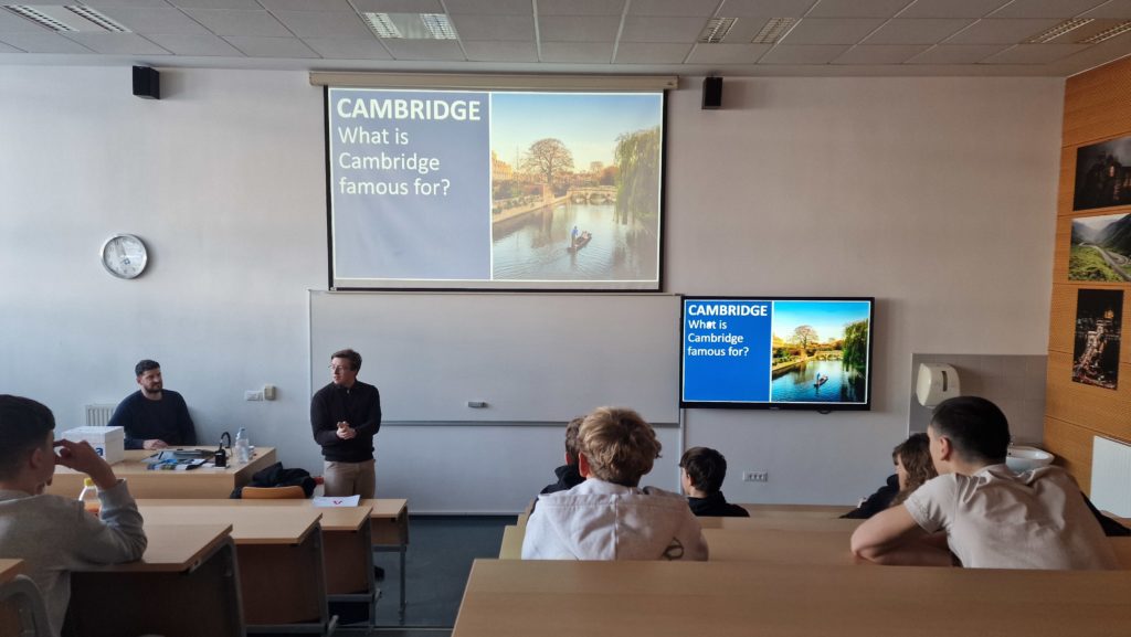 Učilnica na Gimnaziji Šentvid med predstavitvijo jezikovne šole iz Cambridga. Dva predavatelja stojita pred tablo in projektorjem s prosojnico o Cambridgeu, dijaki pa sedijo v klopeh in poslušajo. V ozadju je bela tabla, stenski zaslon in monitor s predstavitvijo.